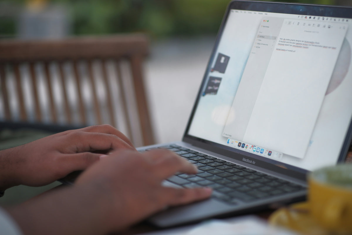 A close-up lifestyle shot of a person typing on a silver MacBook laptop at an outdoor cafe table, with a blurred wooden bench in the background.